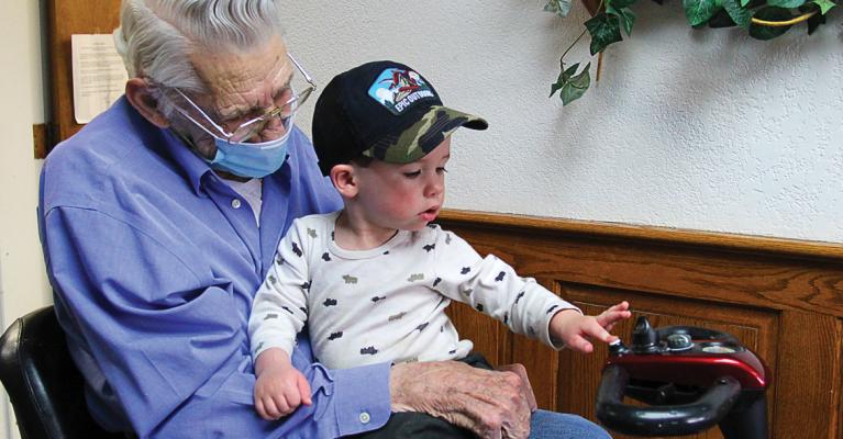 AFTER WAITING for a year, Bill Graff, 90, gets to hold great grandson Von Graff, 1, who presses the scooter’s beeper. Bill is glad for the visit from his family: “Good Lord, it makes life worth living. You betcha.”